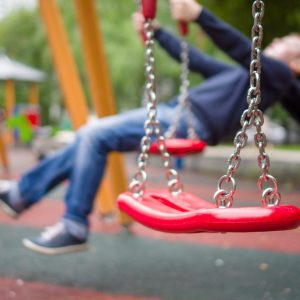 Close-up view of empty red swing with silver chain in front of boy the other swing