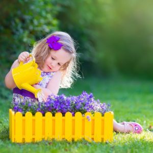 Little girl sits on grass pouring water on summertime flowers from yellow watering pot