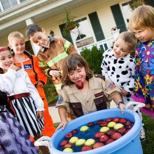 Kid laughs with apple stem in mouth after bobbing for apples with kids nearby in Halloween costumes