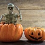 Skeleton sits on top of fake pumpkin next to carved pumpkin and wood planks behind