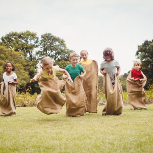 Kids run in burlap sacks across an artificial turf lawn with trees in the background