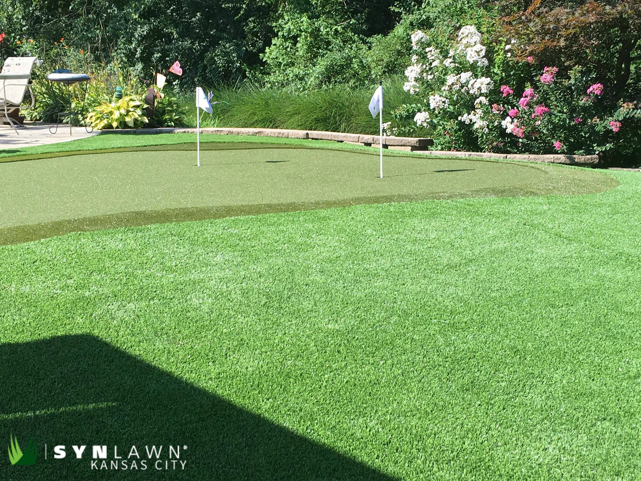 A wide-shot of a synthetic grass putting green with blooming flowers in the background.